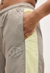 Ręka wsunięta do kieszeni beżowych spodenek z jasnżółtym bocznym panelem i białym logo Champion w pobliżu kieszeni.