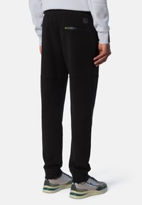 Pantalones de chándal negros con un bolsillo con cremallera, cintura elástica y textura suave, combinados con zapatillas deportivas en gris y verde.
