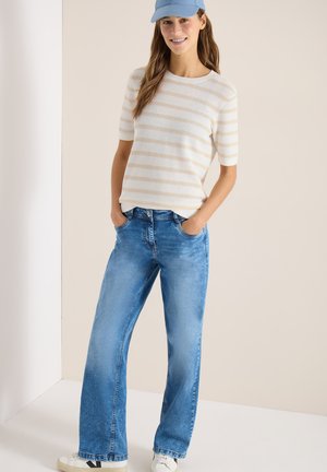 Femme portant un haut beige à rayures à manches courtes, un jean bleu à jambes larges, des baskets blanches et une casquette bleu clair, debout les mains dans les poches.