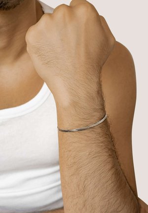 Silver bracelet on a wrist with visible hair; sleek design with a smooth, rounded shape; worn with a plain white sleeveless shirt.
