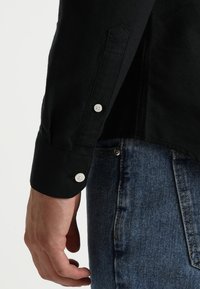 Primer plano de una mano que lleva la manga de una camisa de manga larga negra con botones blancos, descansando cerca del bolsillo de unos jeans de mezclilla azules.