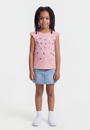 Νεαρό κορίτσι με πλεξούδες, που φοράει ροζ αμάνικη μπλούζα με μπλε γραφικά, τζιν φούστα, λευκές κάλτσες και λευκά παπούτσια slip-on.