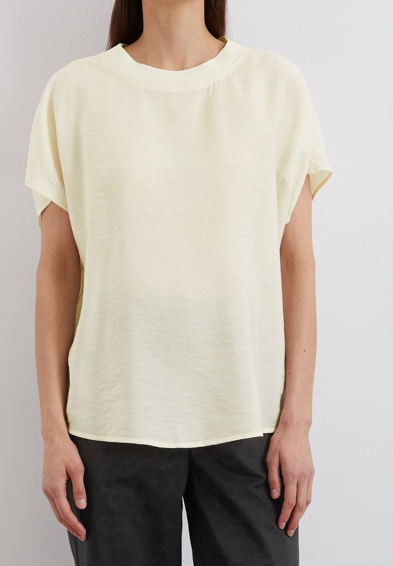 Femme portant une blouse ample à manches courtes de couleur jaune pâle avec un col rond, associée à un pantalon noir, sur un fond uni.