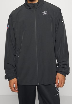 Persoon die een zwarte Raiders jas met rits draagt, met NFL- en Nike-logo's, zichtbaar van schouders tot midden van de dijen tegen een effen achtergrond.