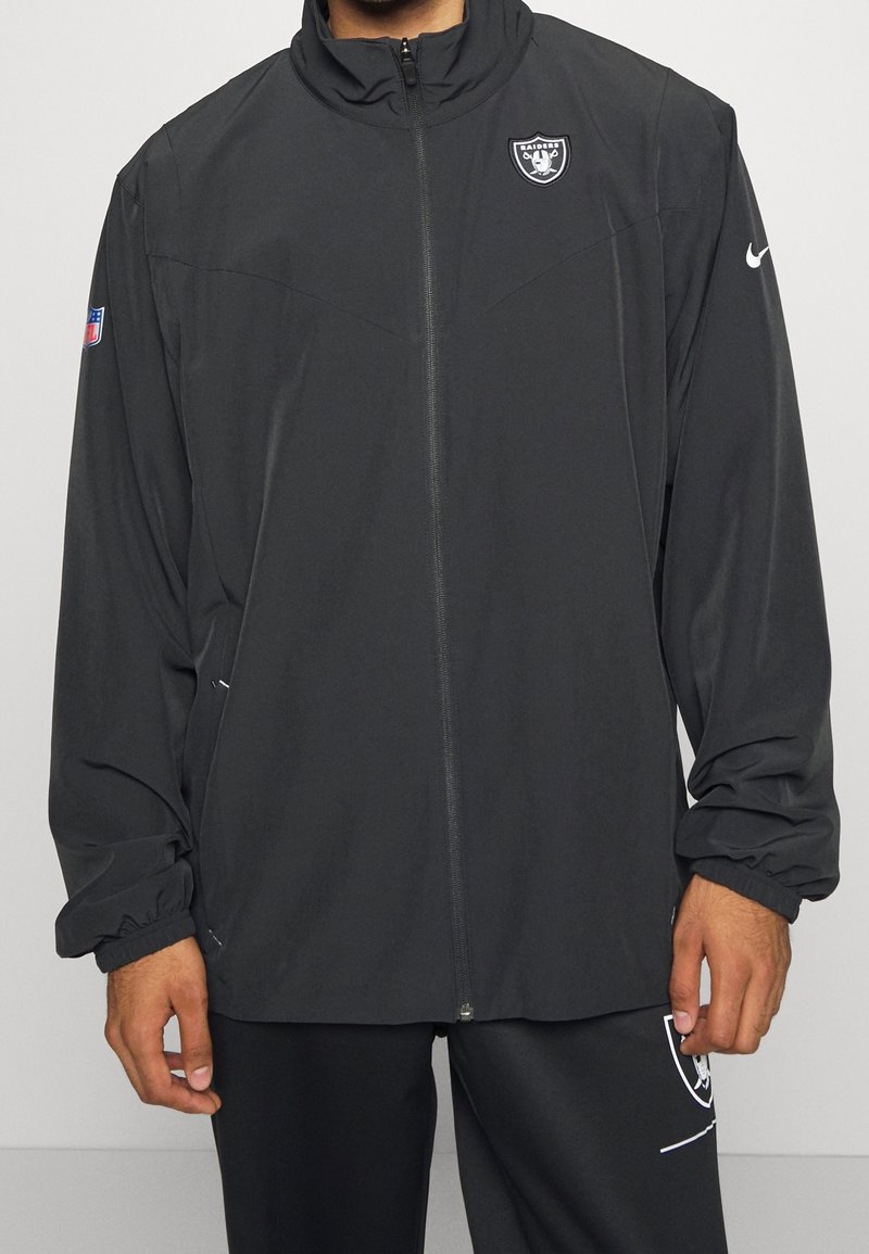 Personne portant une veste Raiders noire zippée avec les logos NFL et Nike, montrée des épaules jusqu'au milieu des cuisses sur fond uni.