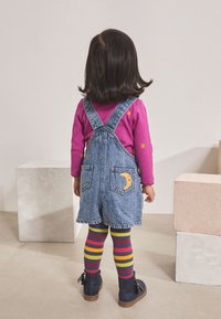 Peto vaquero con parche de luna, camisa morada de manga larga con estrellas, medias de rayas multicolores y botines negros hasta el tobillo.