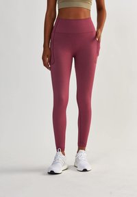 Leggings bordeaux taille haute en tissu extensible avec poches latérales ; associés à des chaussures de sport blanches et un haut beige ajusté.