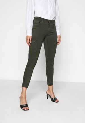 Woman wearing fitted green cargo pants with elastic cuffs, a white long-sleeve shirt, and black open-toe high heel mules.