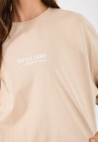 T-shirt de algodão beige com mangas curtas e decote redondo. Apresenta a inscrição "PACIFIC COAST California" impressa em branco na parte da frente.