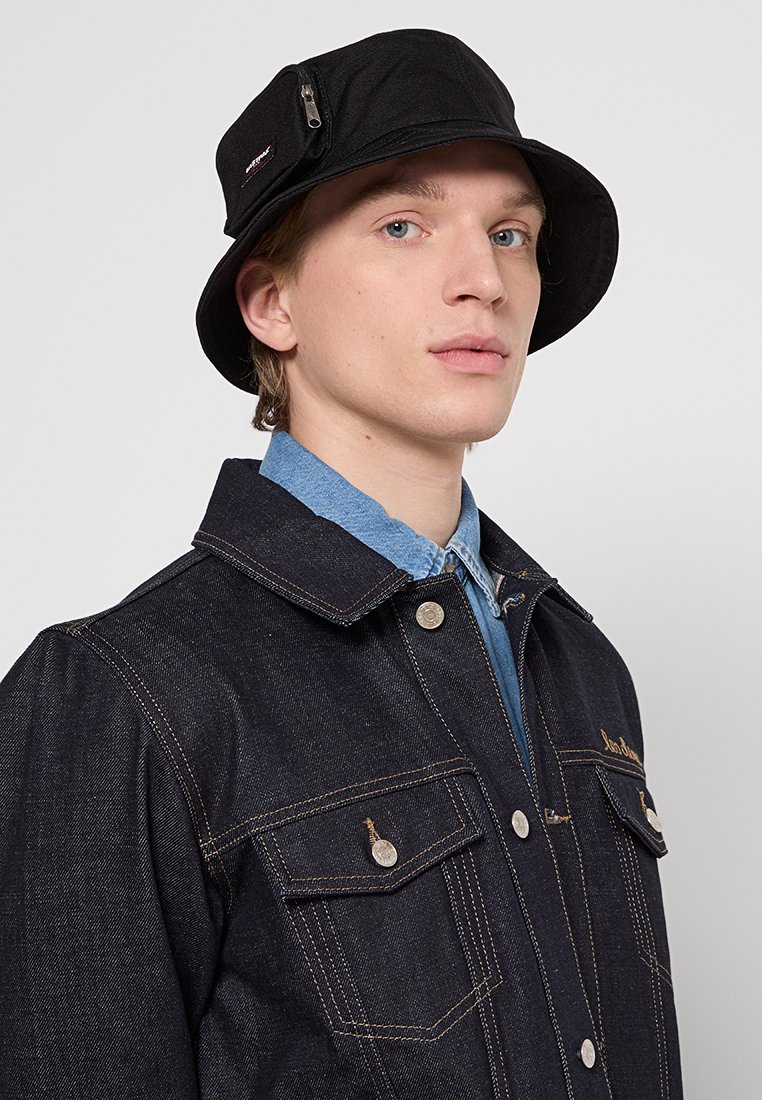 Un jeune homme portant un chapeau seau noir avec une poche zippée, une veste en jean avec des poches à boutons, et une chemise bleue à col, sur un fond uni.