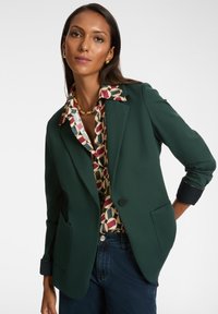 Groene blazer met twee voorkanten zakken, met notch-lapels en contrasterende marineblauwe manchetten. Draagt over een meerkleurig geblokt shirt.