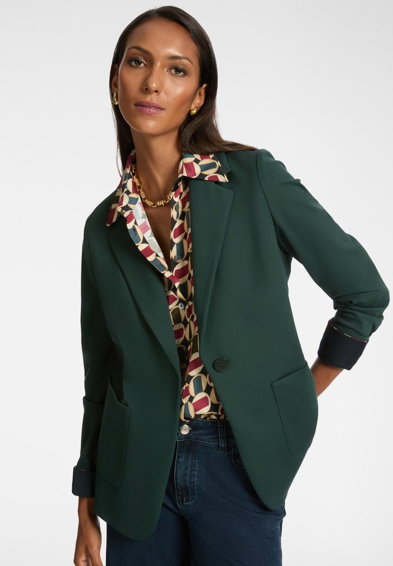 Groene blazer met twee voorkanten zakken, met notch-lapels en contrasterende marineblauwe manchetten. Draagt over een meerkleurig geblokt shirt.