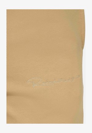 Tissu beige avec une surface texturée présentant un léger relief. Logo brodé blanc dans le coin inférieur. Détail en gros plan.