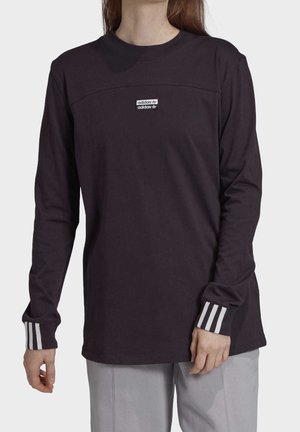 Personne portant un t-shirt Adidas noir à manches longues avec le logo sur la poitrine et trois rayures blanches aux poignets, associé à un pantalon gris clair.