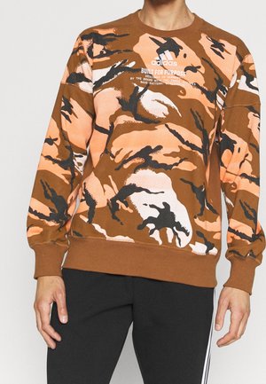 Homme portant un sweat Adidas à motif camouflage marron, orange, blanc et noir avec le texte "Built for Purpose" et un pantalon noir à rayures blanches.