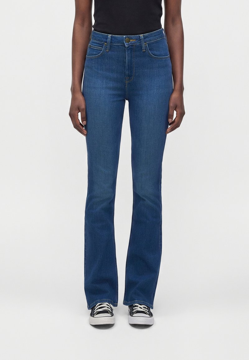 Persoon draagt donkerblauwe bootcut jeans met zwart-witte sneakers, staand tegen een effen lichte achtergrond.
