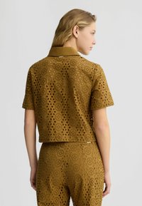 Kurzarmpullover aus braunem, durchlöchertem Stoff mit kreisförmigen Ausschnitten und einem klassischen Kragen, kombiniert mit passenden hochgeschnittenen Hosen.