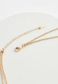 Collier en chaîne à plusieurs couches de couleur or, doté d'un fermoir à mousqueton, d'un design en maillons plats et d'une chaîne d'extension ajustable avec une petite étiquette.