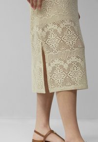 Knielange Strickrock in Creme mit aufwendigen Durchbruchs-Mustern und einem seitlichen Schlitz. Das Material wirkt texturiert und hat ein leichtes Gefühl.