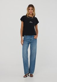 T-shirt nera a maniche corte con dettagli di borchie e testo ricamato, abbinata a jeans a gamba larga di un blu chiaro e sandali con tacco alto.