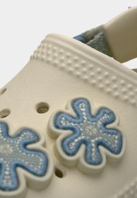 Gros plan d'un sabot en caoutchouc beige avec un bord texturé et deux breloques décoratives en forme de fleur bleue attachées.