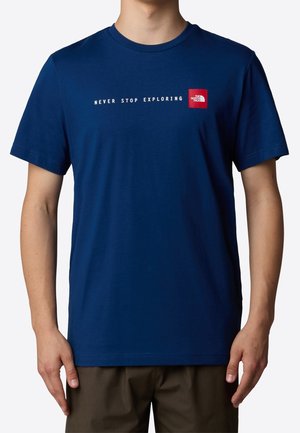 Mężczyzna w niebieskiej koszulce z krótkim rękawem z białym napisem "NEVER STOP EXPLORING" i czerwonym logo The North Face na piersi.