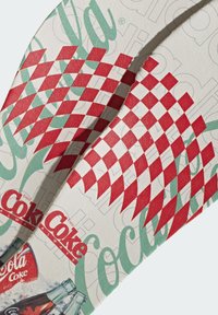 Tongs blanches avec motif de drapeau à damier rouge et texte vert « Coca-Cola », avec des illustrations vintages de bouteilles de Coca-Cola sur la semelle.