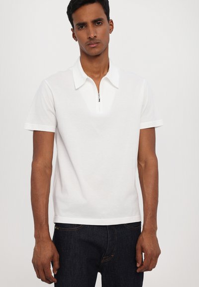 Balta polo marškinėliai su užtrauktuku priekyje, trumpomis rankovėmis ir kontrastine apykakle. Lygus medvilnės audinys, pritaikyta išvaizda, derinama su tamsiomis džinsais.