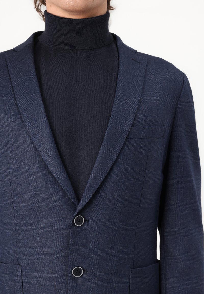 Homme portant un blazer à carreaux bleu marine foncé sur un pull à col roulé noir, montré de la nuque à la taille sur un fond blanc.