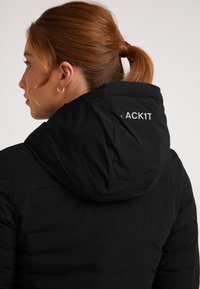 Sort isoleret jakke med hætte, der har teksten "JACK1T" i reflekterende sølv på ryggen. Glat tekstur med et strømlinet design.
