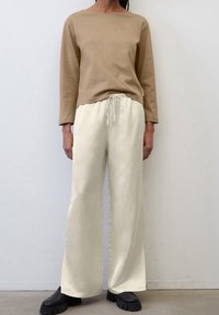 Beige langärmeliger Pullover mit einem Bootsausschnitt, kombiniert mit cremefarbenen, weit geschnittenen Hosen mit Kordelzug, beide aus glattem Stoff. Schwarze klobige Stiefel.