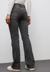 I jeans neri a zampa di elefante presentano una finitura testurizzata, un design a cinque tasche e cuciture in vista, con vita a media altezza. Indossati con scarpe di colore chiaro.