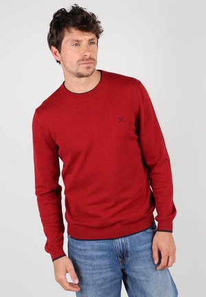 Pull rouge en tissu tricoté, avec un col rond, des manches longues et un ourlet côtelé. Détail subtil du logo sur la poitrine. Porté avec un jean bleu.