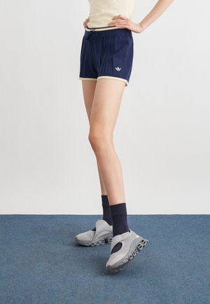Person trägt marineblaue Sportshorts mit beigen Akzenten, schwarze Socken und graue Sportschuhe und steht auf blauem Teppich.