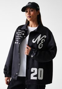 Black satin coach jacket featuring a white "NE" logo, silver lettering, and number "20". Paired with a black cap and white T-shirt.