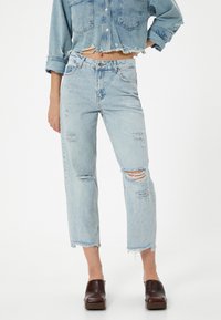 Vaqueros de denim de color azul claro, desgastados, con corte recto, dobladillo deshilachado y múltiples rasgaduras. Combinados con una chaqueta de denim corta a juego.