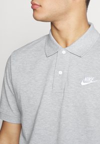 Man som bär en ljusgrå kortärmad Nike-polo med vitt broderat logo, som tittar åt sidan mot en enfärgad bakgrund.