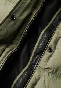 Gros plan sur une veste kaki ouverte avec une fermeture éclair noire et des boutons pression, montrant la doublure intérieure noire et le ruban de marque le long du bord de la fermeture éclair.
