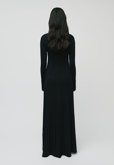 Donna con lunghi capelli ondulati scuri, vestita con un abito nero a coste lungo fino ai piedi e con maniche lunghe, in piedi con le spalle rivolte verso di noi su uno sfondo neutro.
