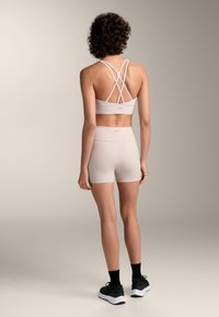 Svetloružový športový podprsenka s krížovými ramienkami a zodpovedajúcimi šortkami s vysokým pásom. Mäkka textúra, priliehavý dizajn a minimálne logo na zadnej strane.