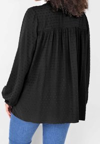 Blusa nera, текстurizzata con un sottile motivo a pois, maniche lunghe e schiena arricciata, abbinata a jeans azzurri chiari.