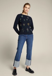 Marineblauwe trui met bloemenborduursel, gecombineerd met lichte spijkerbroeken met een contrasterende zoom. Zwarte loafers maken de outfit compleet.
