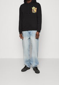 Haut à capuche noir avec un graphique sur la poitrine, associé à un jean large bleu clair. Les chaussures sont noires avec une surface texturée.