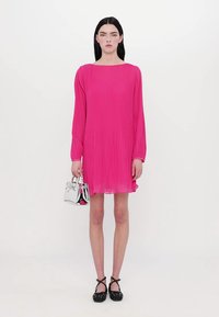 Roze langwerpige geplooide jurk, knie-lang, met een vierkante halslijn. Model houdt een kleine zilveren handtas vast en draagt zwarte schoenen met stippen.