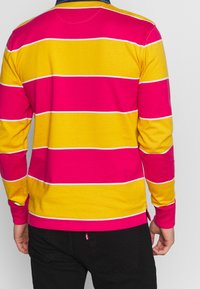 Långärmad polotröja med breda rosa och gula ränder, tillverkad av mjukt tyg med ribbad krage och en liten logotagg vid fållen.