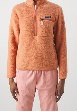 Person iført en ferskenfarvet sherpa fleecepullover med lynlås på brystlommen og lyserøde bukser, stående foran en ensfarvet baggrund.