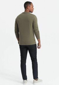 Camisa de manga larga verde oliva con escote redondo, combinada con vaqueros oscuros y zapatillas blancas, vista desde atrás.