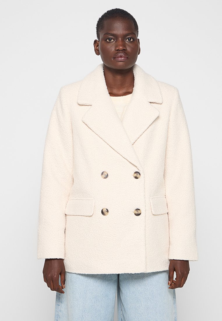 Czarnoskóra kobieta ubrana w kremowy, dwurzędowy, teksturowany płaszcz wełniany z dużymi klapami i jasnoniebieskie dżinsy, stojąca na jednolitym tle.