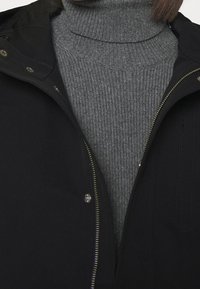 Black outer jacket with a zip and snap closure, layered over a textured grey turtleneck sweater featuring ribbed patterns.