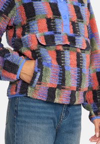 Tekstureret fleece pullover med et patchwork-design i rød, grøn, blå og sort. Har en knappet hals og en frontlomme.